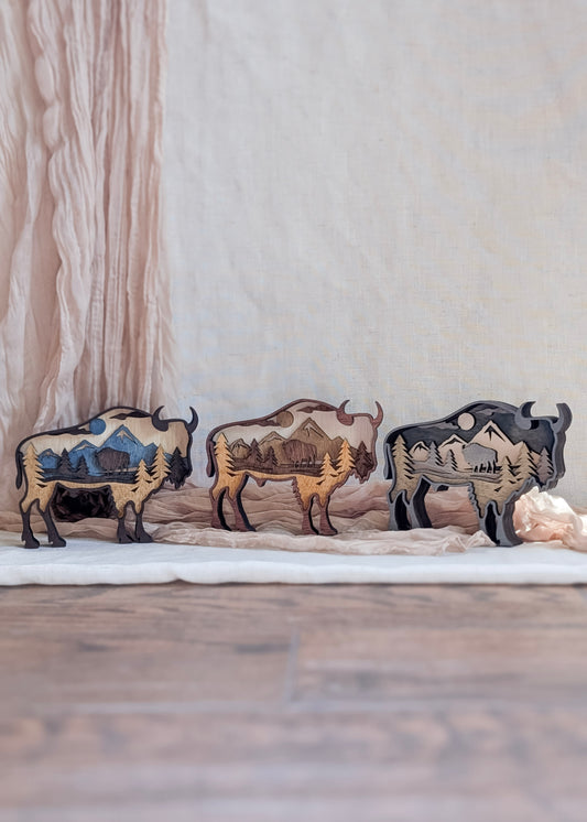 Three layered bison wood art pieces shown side by side, displaying stain color options: rustic brown, gray wash, and neutral natural wood.