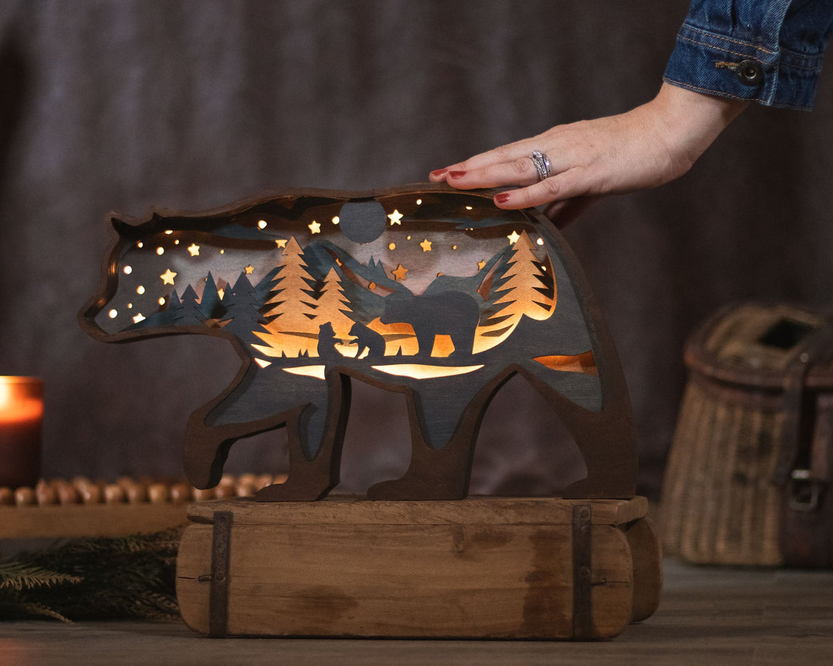 A bear-shaped wooden night light with a cutout forest and mountain scene, glowing with warm light, displayed on a rustic wooden base. A person's hand is resting on the bear's back, and a candle and woven basket are softly blurred in the background.