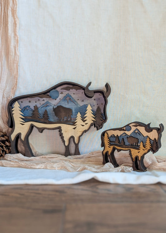 Layered laser-cut wood art showing a 3D bison silhouette with mountains, pine trees, and a moonlit night scene.