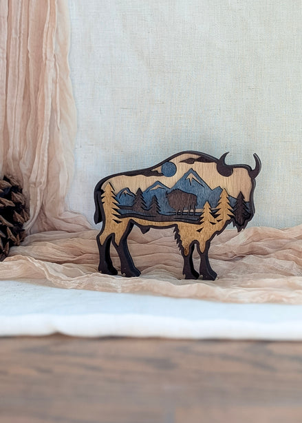 Laser-cut bison wood art in rustic brown stain with layered mountain scene and blue sky detail.