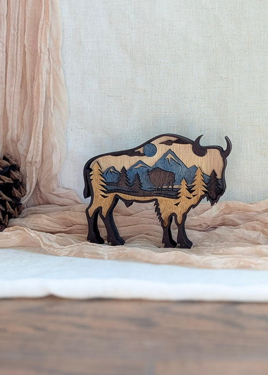 Laser-cut bison wood art in rustic brown stain with layered mountain scene and blue sky detail.