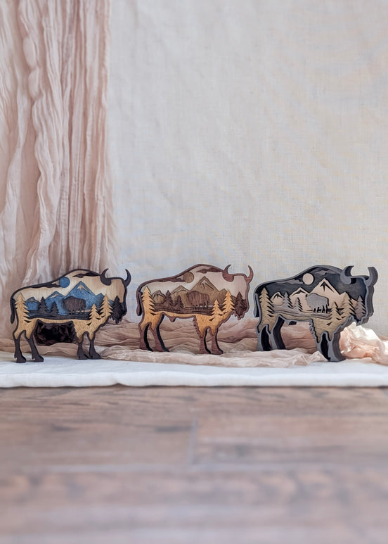 Three layered bison wood art pieces shown side by side, displaying stain color options: rustic brown, gray wash, and neutral natural wood.