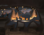 A wooden bear-shaped night light with a laser-cut forest and mountain scene, glowing warmly from internal lighting. The design includes a bear silhouette, pine trees, stars, and mountains shown against a dark background.