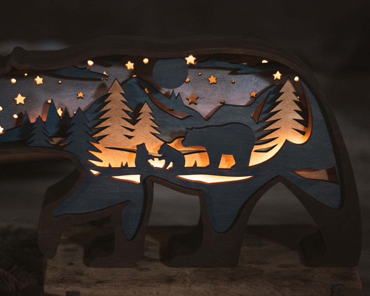 A wooden bear-shaped night light with a laser-cut forest and mountain scene, glowing warmly from internal lighting. The design includes a bear silhouette, pine trees, stars, and mountains shown against a dark background.