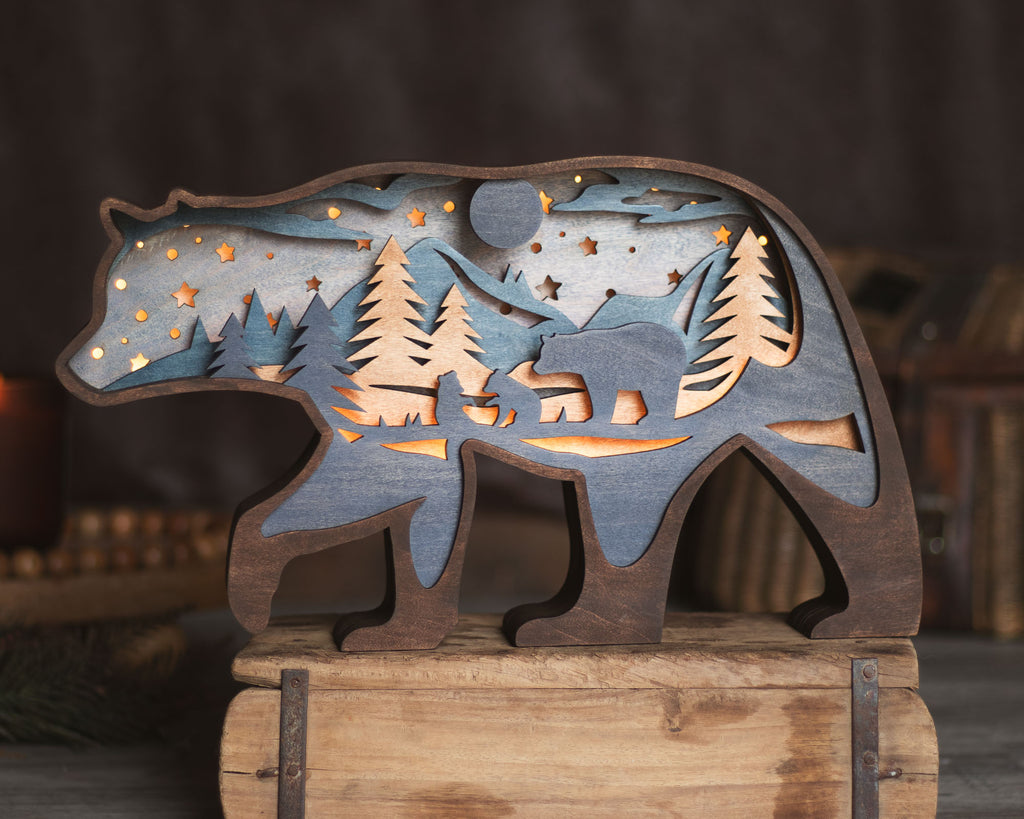 A rustic wooden bear-shaped decorative piece with layered cutout details showing a forest, moon, stars, and a smaller bear silhouette, displayed on a wooden crate against a softly blurred dark background.