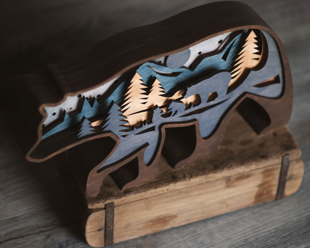 A rustic wooden cutout sculpture of a bear silhouette layered with a mountain and pine forest scene, displayed on a wooden stand against a dark wood background.
