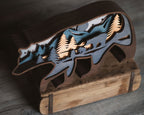 A rustic wooden cutout sculpture of a bear silhouette layered with a mountain and pine forest scene, displayed on a wooden stand against a dark wood background.