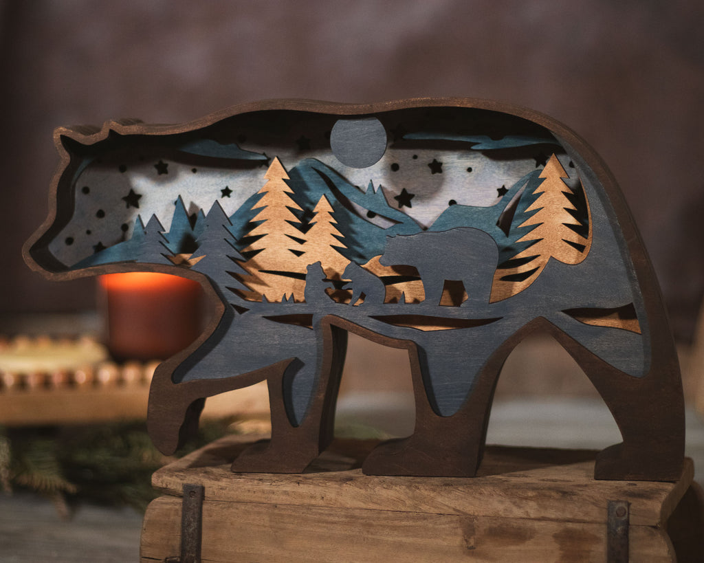 A rustic wooden bear-shaped decorative sculpture with layered cutout details of mountains, pine trees, stars, and a bear silhouette, displayed on a wooden surface with a warm blurred background.
