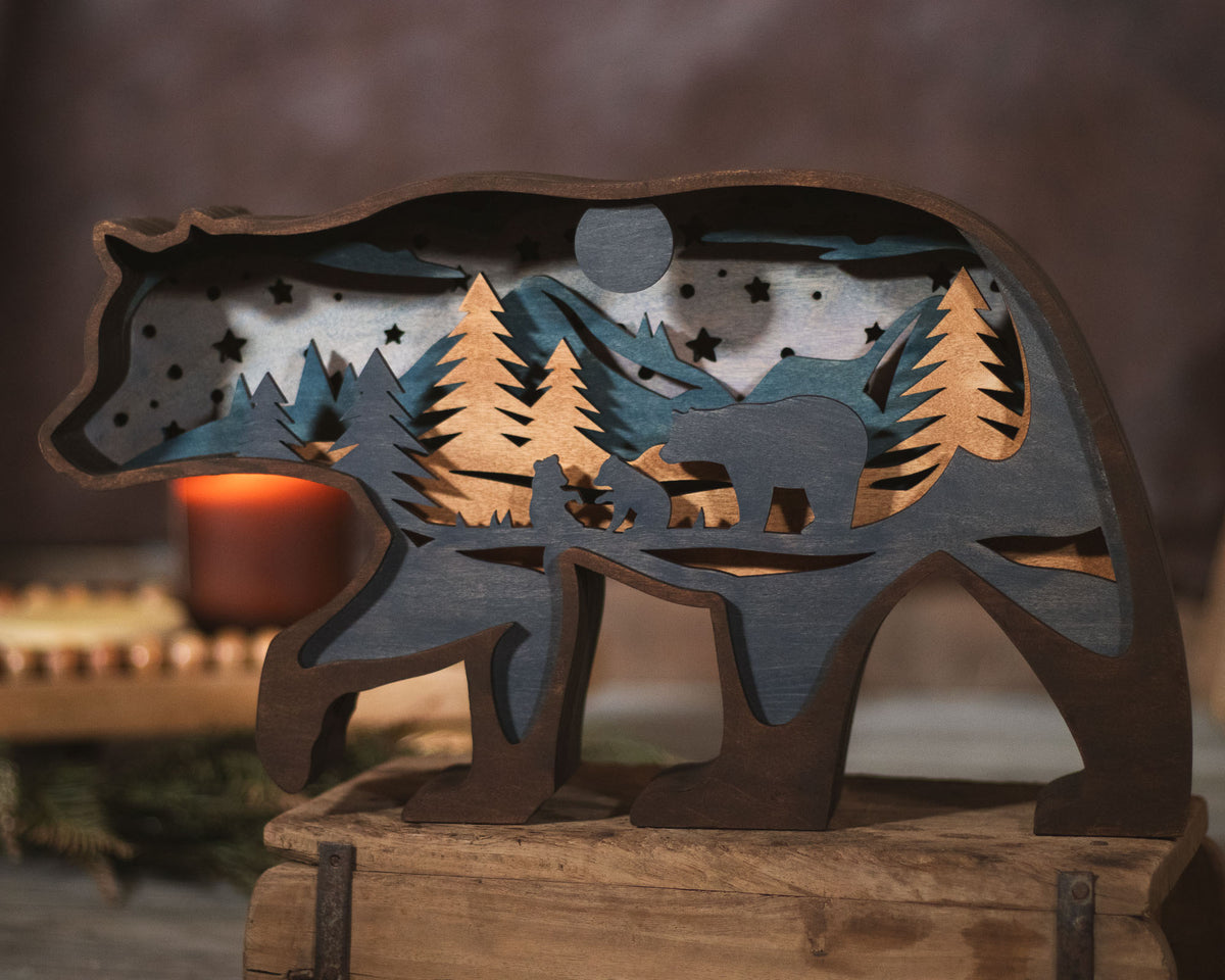 A rustic wooden bear-shaped decorative sculpture with layered cutout details of mountains, pine trees, stars, and a bear silhouette, displayed on a wooden surface with a warm blurred background.