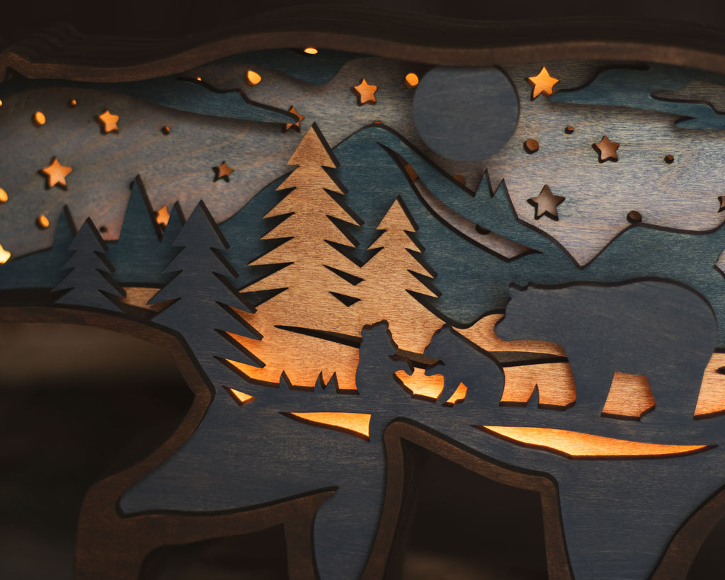 Close-up of a laser-cut wooden night scene with silhouettes of pine trees, mountains, stars, a bear, and a deer, lit from within with warm amber light.