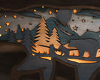 Close-up of a laser-cut wooden night scene with silhouettes of pine trees, mountains, stars, a bear, and a deer, lit from within with warm amber light.