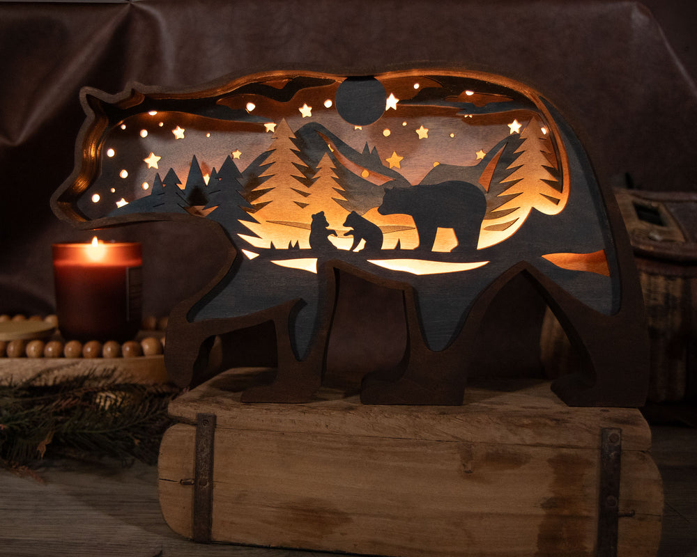 A carved wooden bear-shaped night light with a mother bear and two baby bears, trees, mountains, and stars cut out, glowing warmly from within. It is displayed on a rustic wooden box with a lit candle and cozy decor in the background.