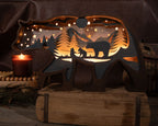 A carved wooden bear-shaped night light with a mother bear and two baby bears, trees, mountains, and stars cut out, glowing warmly from within. It is displayed on a rustic wooden box with a lit candle and cozy decor in the background.