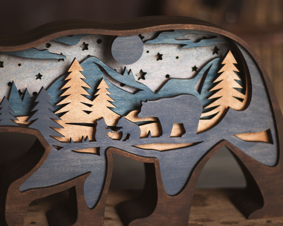 A close-up of a layered wooden bear-shaped decorative piece with a night landscape design, featuring pine trees, mountains, stars, and a bear silhouette in shades of natural wood and weathered blue-gray.