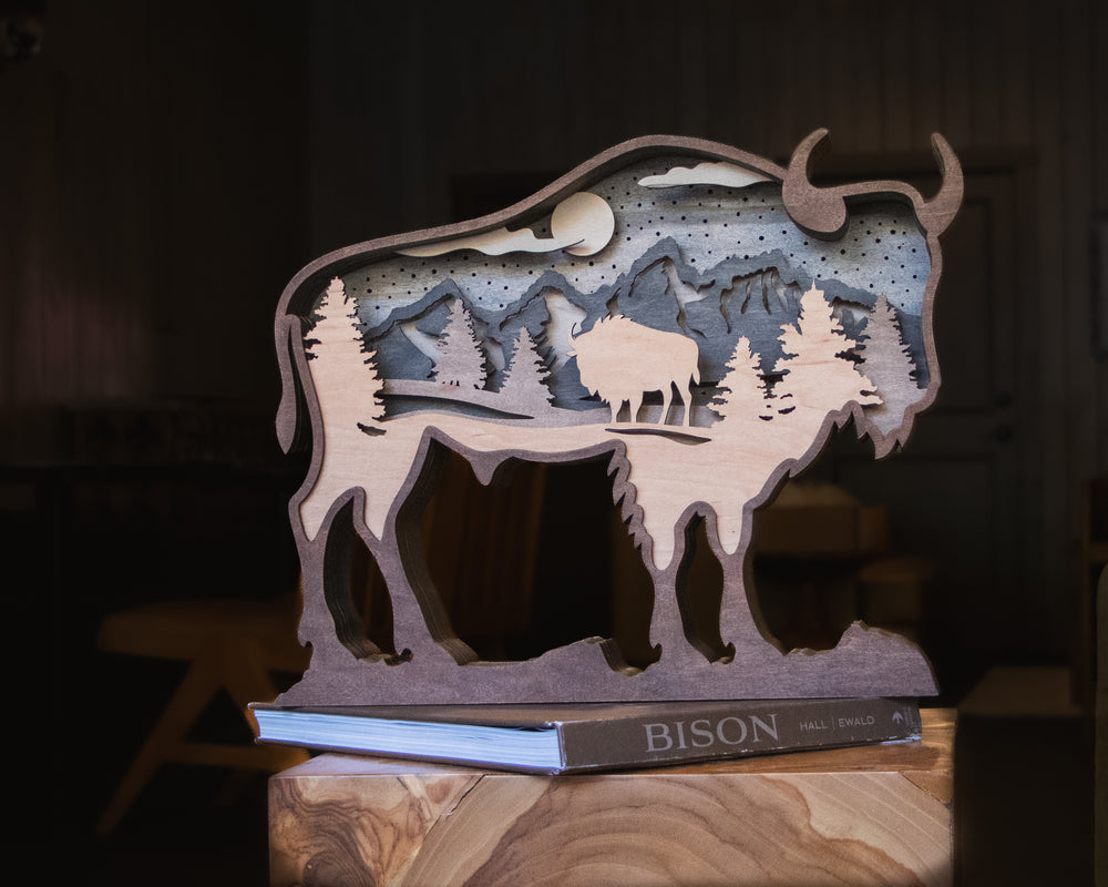 A laser-cut wooden bison silhouette sculpture with layered mountain, tree, and moon details, displayed on a book resting on a wooden surface in a dim indoor setting.