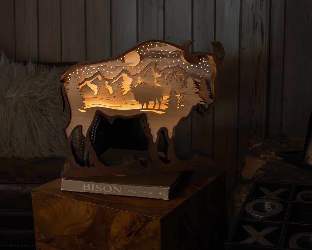 A lit wooden bison-shaped decorative sculpture with a mountain scene, displayed on a book on a wooden side table in a rustic room.