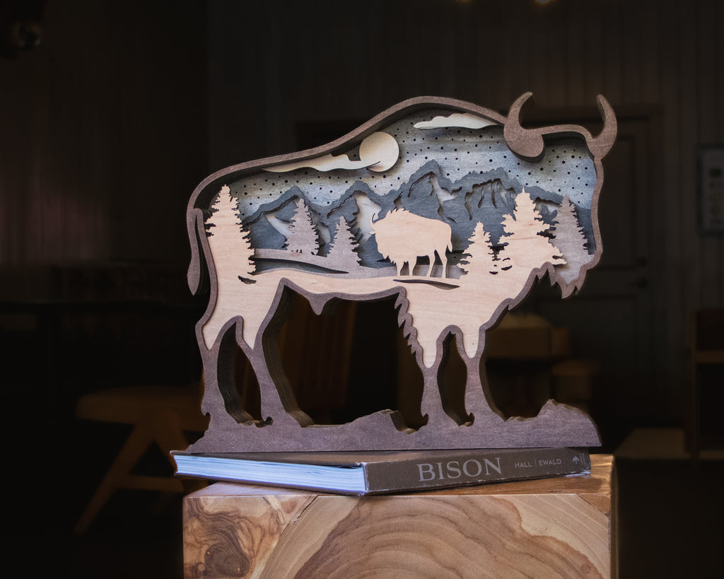 A layered wooden cutout sculpture of a bison silhouette containing a mountain and tree landscape scene, displayed on a book labeled BISON in a dimly lit indoor setting.