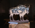 A layered wooden cutout sculpture of a bison silhouette containing a mountain and tree landscape scene, displayed on a book labeled BISON in a dimly lit indoor setting.