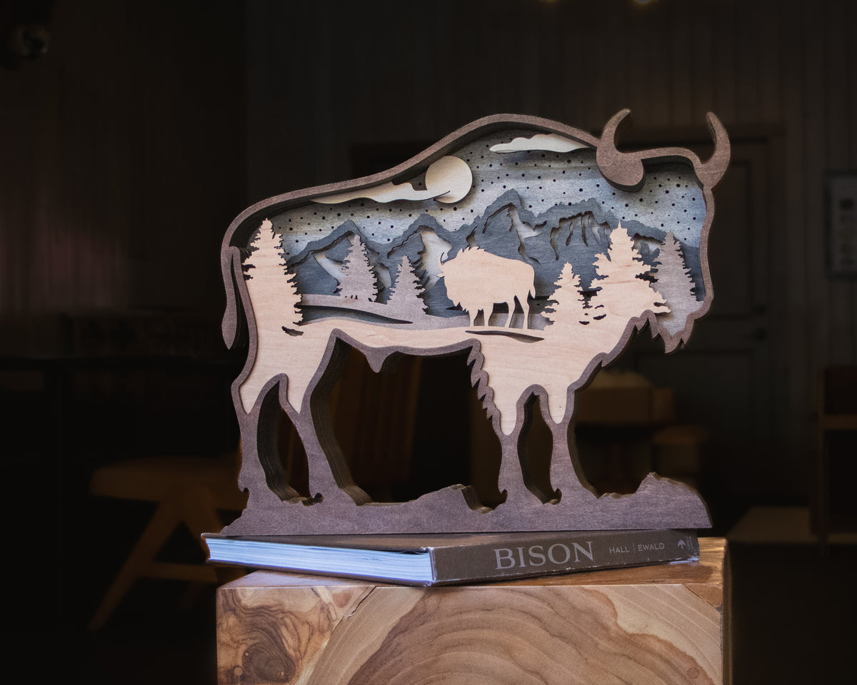 A layered wooden cutout sculpture of a bison silhouette containing a mountain and tree landscape scene, displayed on a book labeled BISON in a dimly lit indoor setting.