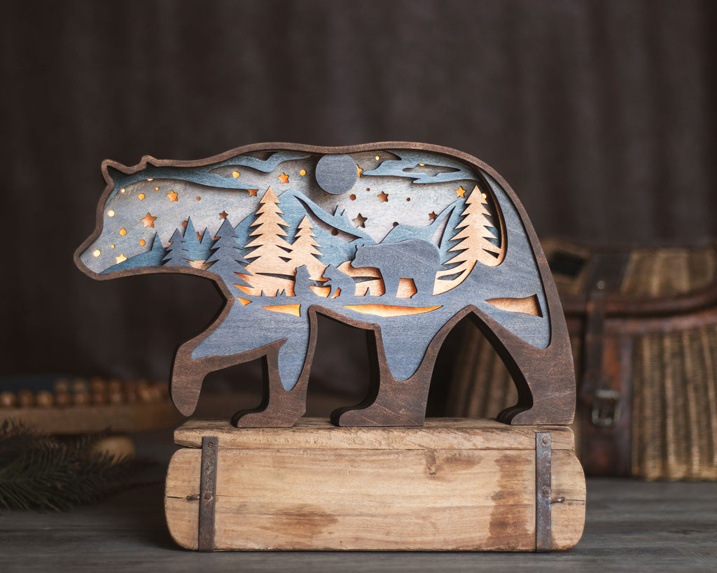 A wooden bear-shaped decorative light with laser-cut forest and starry night details, glowing from within, displayed on a rustic wooden base against a dark, blurred background.