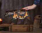 A bear-shaped wooden night light with a cutout forest and mountain scene, glowing with warm light, displayed on a rustic wooden base. A person's hand is resting on the bear's back, and a candle and woven basket are softly blurred in the background.