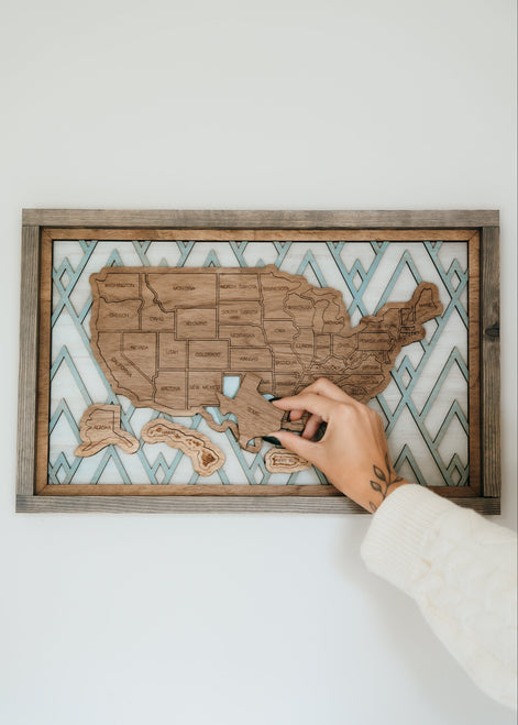 Hand placing a wooden state piece onto the Summit Peaks travel map from the Storyline Series, featuring a mountain-inspired background.